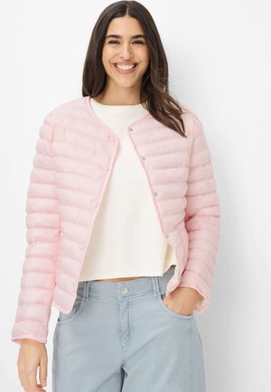 Lächelnde Frau trägt eine hellrosa gesteppte Jacke über einem weißen Top und hellblauen Hosen, mit einer Hand in der Tasche vor einem weißen Hintergrund.