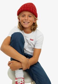 Biely tričko s červeným logom Levi's, tmavomodré džínsy, červená pletená čiapka, biele ponožky, červené tenisky Converse, sediaci na bielom stoličke.
