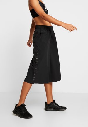 Frau trägt schwarze weite Under Armour Capri-Hosen, schwarzen Sport-BH und schwarze Laufschuhe, steht seitlich mit einem ausgestreckten Arm.