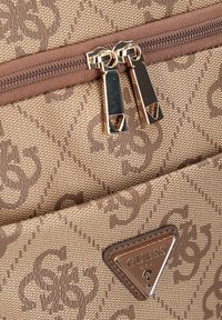 Rucsac textil maro cu o suprafață cu model, prevăzut cu fermoare aurii, o plăcuță logo triunghiulară și o textură distinctă.