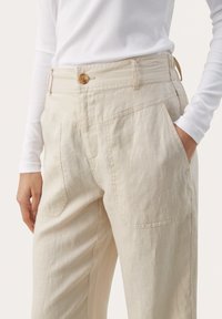 Beige linen trousers with a smooth texture, featuring a button closure, side pockets, and a relaxed fit. Tucked into a fitted white long sleeve top.