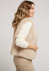 Gilet en sherpa beige avec une surface douce et texturée, sans manches, et une coupe ample, associé à un haut à manches longues crème et un pantalon beige.