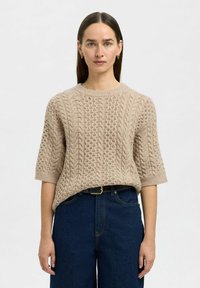 Suéter beige de punto con un patrón de cable texturizado, mangas cortas y un escote redondeado, combinado con unos jeans de tiro alto en azul oscuro.