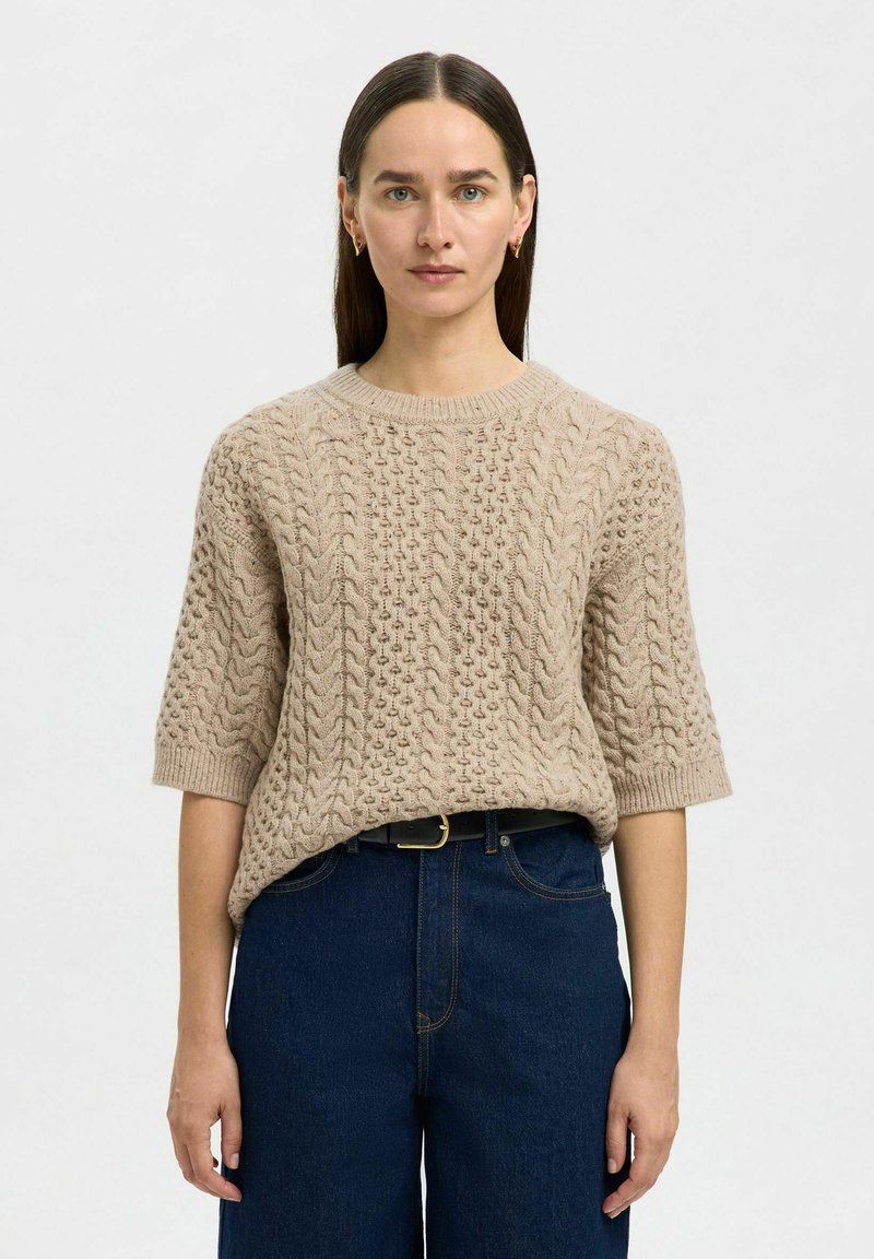 Suéter beige de punto con un patrón de cable texturizado, mangas cortas y un escote redondeado, combinado con unos jeans de tiro alto en azul oscuro.