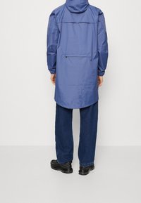 Blå, letvægts, vandtæt jakke med hætte, lange ærmer og en baglomme, kombineret med løstsiddende mørke jeans og sorte sko.