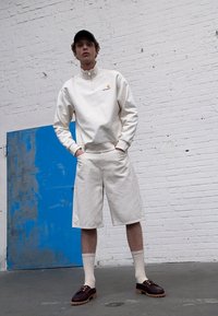 Witte zip-up sweatshirt met logo, beige shorts, witte sokken en donkere schoenen met gestructureerde zolen. De achtergrond heeft een blauwe muur en bakstenen.