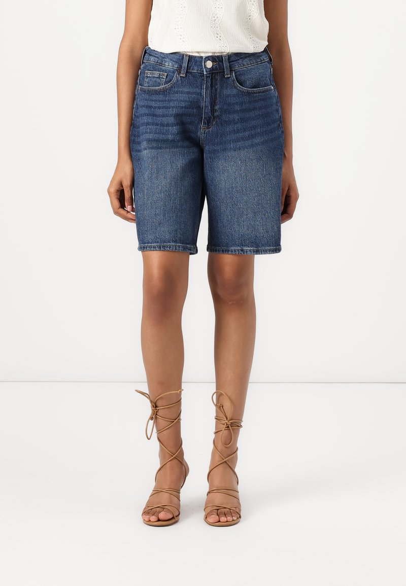 Bermudas de mezclilla con un lavado medio, estilo de cinco bolsillos y un corte recto. Combinadas con sandalias de tiras de color marrón claro.