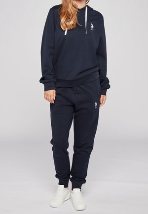 Vrouw die een zwart hoodie en bijpassende trainingsbroek draagt met witte logo's en witte sneakers, staand tegen een effen achtergrond.