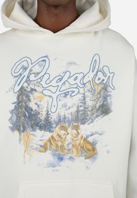 Cremefarbener Hoodie mit einer Grafik von zwei Wölfen vor einem schneebedeckten Hintergrund, mit blauer Schrift darüber und einer Fronttasche an der Basis.