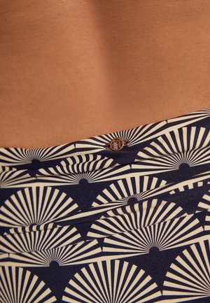 Közeli kép mintás tengerészkék és fehér anyagról, rajta legyező alakú geometriai formák és egy kis kerek fém embléma mezítelen bőrön.