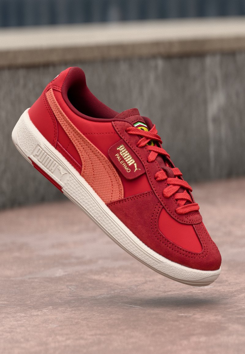 Sneaker rouge en alliant des matériaux en daim et en mesh, avec une semelle blanche texturée et une marque dorée bien visible sur le côté.