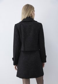 Žena s dlouhými blond vlasy v černém krátkém saka s drobnými skvrnkami a odpovídající sukní, stojící zády k světlému šedému pozadí.