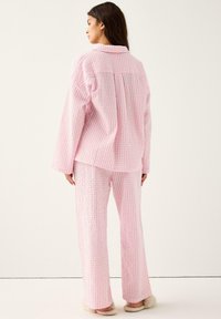 Donna di spalle che indossa un completo pigiama a maniche lunghe a scacchi rosa e bianchi, con pantaloni abbinati e pantofole fuzzy beige.