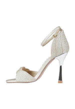 Sandalo con tacco alto argentato glitterato con cinturino alla caviglia, punta aperta e tacco stiletto metallizzato lucido su sfondo bianco.