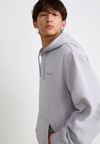 Haut à capuche gris clair en tissu doux. Il possède une poche avant, un capuchon avec cordon de serrage ajustable et un logo brodé discret sur la poitrine.