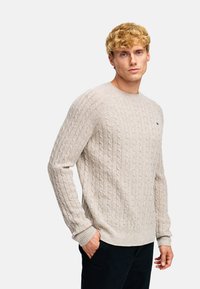 Jasno beżowy sweter o splocie w warkocze z długimi rękawami, ściągaczami u mankietów i na dole. Posiada małe logo na piersi oraz klasyczny dekolt typu crew neck.