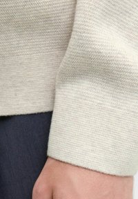 Hellbeiger Strickpullover aus glattem Material. Verfügt über ein Rippbündchen und ein figurbetontes Design, das teilweise einen dunklen Stoff darunter zeigt.