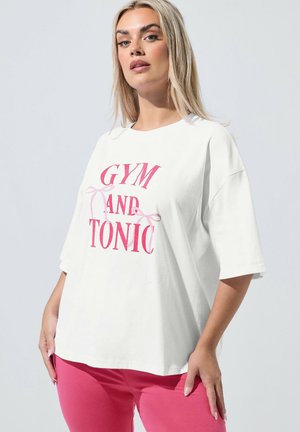 Mujer rubia con una camiseta blanca de gran tamaño con el texto "GYM AND TONIC" en rosa y pantalones rosa brillante, de pie contra un fondo claro.