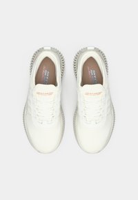 White mesh sneakers with textured rubber soles, featuring a lace-up design and branding on the tongue. Accents in light gray and peach.