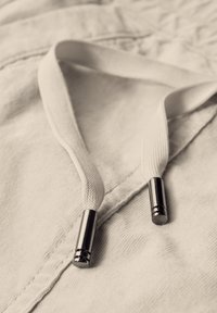 Cordon en tissu blanc avec des embouts métalliques, reposant sur une surface texturée claire. Les embouts ont une finition brillante et une forme cylindrique.