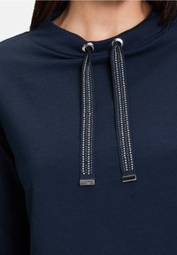 Sudadera azul marino con cuello redondo, con cordones decorativos brillantes y acentos en metal plateado. Textura de tela suave.