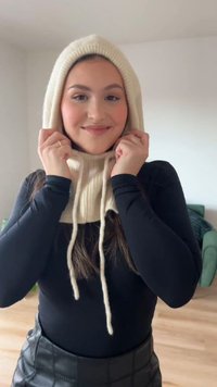 Kremowa dzianinowa bluza z kapturem o prążkowanej fakturze i sznurkami do ściągania. Noszona z czarną bluzką z długim rękawem. Jasne tło z zielonym meblem.