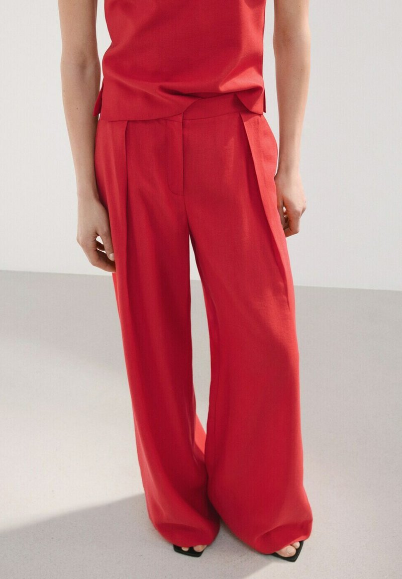 Person trägt lockere, hoch taillierte rote Hose kombiniert mit einem passenden roten Oberteil, steht auf einem hellgrauen Boden.