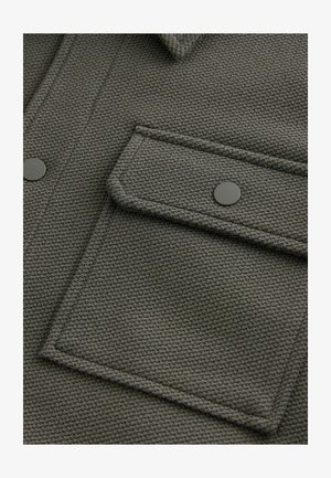 Roheline tekstureeritud kangas, millel on klapiga tasku ümarate servadega ja musta nupu tõmbega, samuti peene kootud mustriga kogu pinna ulatuses.