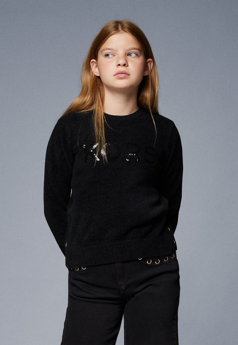 Maglione lavorato a maglia nero con lettere in paillettes e occhielli dorati decorativi all'orlo, abbinato a pantaloni neri aderenti.