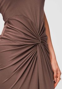 Gros plan d'une robe marron avec un tissu froncé torsadé à la taille, montrant une texture lisse et une main avec des bagues reposant sur le côté.