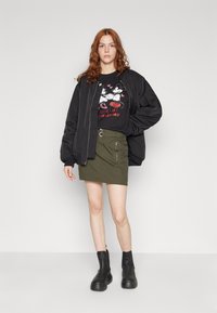 Fekete bomber dzseki, grafikai mintás póló Mickey Mouse dizájnnal, olíva zöld miniszoknya, térd fölé érő fekete csizma tömör talppal.