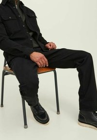 Chaqueta de camisa negra con dos bolsillos en el pecho sobre una blusa verde oscuro; combinada con pantalones negros a juego y zapatos negros con cordones.