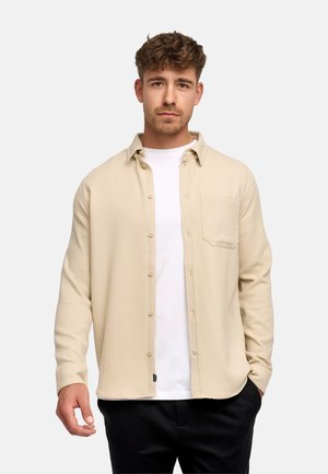 Uomo con capelli corti e ricci che indossa una camicia beige con bottoni sopra una t-shirt bianca e pantaloni neri, in piedi davanti a uno sfondo semplice.