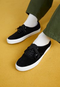 Mustad korgitsneakerid valge kummist tallaga, millel on paelad ja väikesed aasad, mille juurde on sobitatud valged lühikesed sokid ja rohelised püksid.