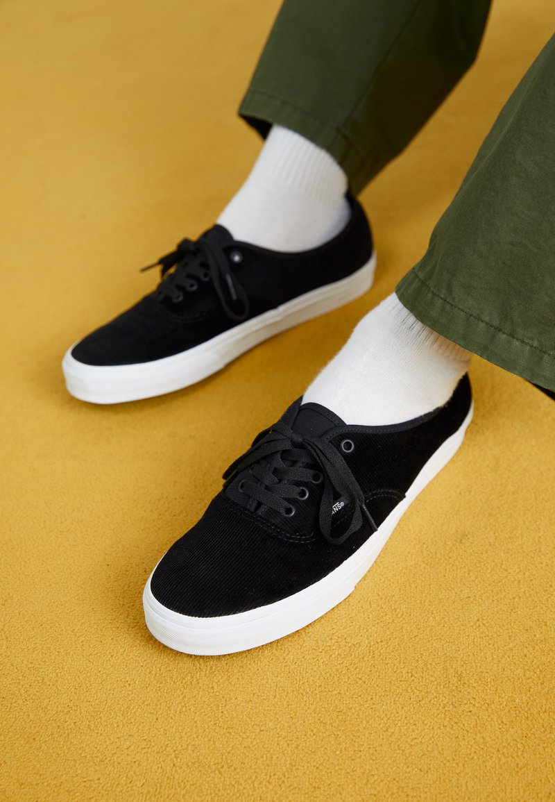 Mustad korgitsneakerid valge kummist tallaga, millel on paelad ja väikesed aasad, mille juurde on sobitatud valged lühikesed sokid ja rohelised püksid.