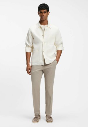 Man staat en draagt een crèmekleurig jasje met knopen over een wit overhemd, beige broek en beige loafers met kwastjes tegen een effen witte achtergrond.