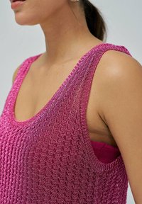 Žena nosi bezrukavu, teksturiranu ružičastu pletenu majicu preko tamije ružičaste donje odjeće, prikazana od vrata do ramena na jednostavnoj pozadini.