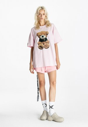 Mujer rubia con camiseta rosa oversize con gráfico de osito de peluche, pantalones cortos rosas a juego, calcetines blancos con diseño negro y zapatos beige texturizados.