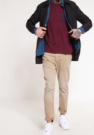 Mies, jolla on musta takki sinisen ruudullisen paidan ja viininpunaisen t-paidan päällä, beigeihin käännettyihin housuihin pukeutuneena, valkoiset lenkkarit jalassa ja tatuoitu käsi korjaa takkia.