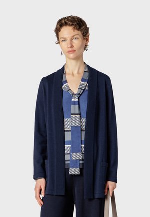 Femme portant un cardigan bleu marine et une écharpe bleue à motifs géométriques sur un haut bleu, tenant un sac cabas beige devant un fond uni.