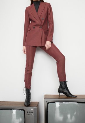 Mujer vestida con un traje rojo de cuadros, suéter de cuello alto negro y botas negras de tacón, de pie con un pie sobre dos televisores vintage.