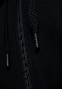 Primo piano della cerniera di un hoodie nero e delle corde di regolazione intrecciate con il nome del marchio Lacoste sulle estremità in plastica.