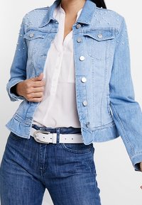 Ljust blå denimjacka med silvernitar på axlarna, klassisk krage och fickor fram. Har metallknappar och en figurnära design.