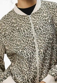 Veste bomber à imprimé léopard en beige, avec une fermeture éclair frontale, un col et des poignets côtelés, présentant une texture lisse et un motif distinct.