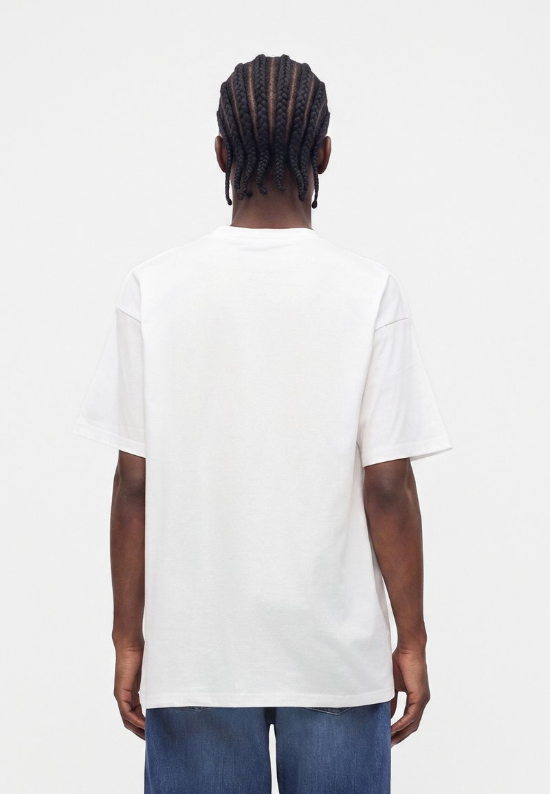 Person mit geflochtenem Haar, die ein schlichtes weißes kurzärmeliges T-Shirt und blaue Jeans trägt, von hinten vor weißem Hintergrund gesehen.