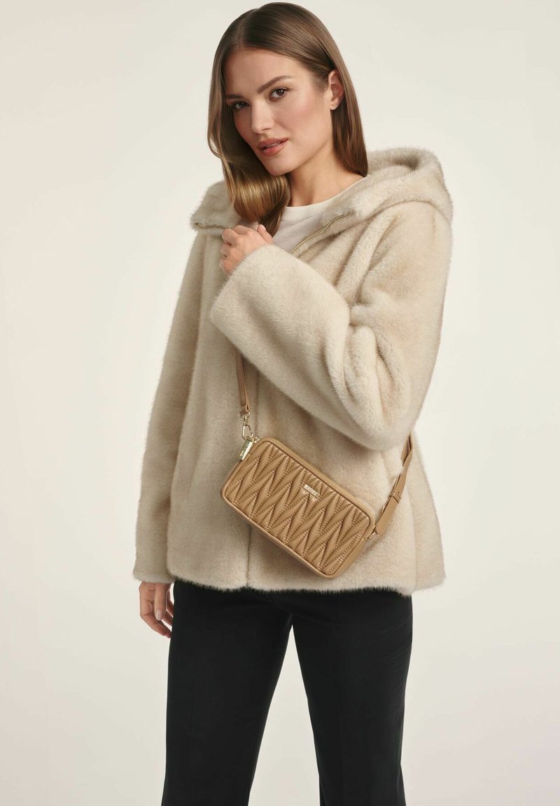 Beżowa kurtka z faux futra z kapturem, w połączeniu z brązową pikowaną torbą na ramię. Widoczne czarne spodnie, ukazujące miękki, teksturowany wygląd.