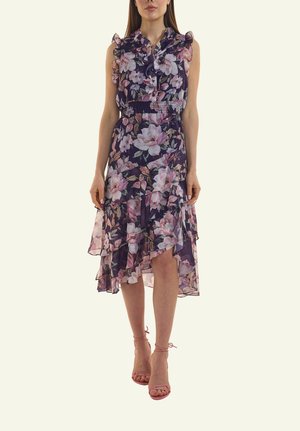 Frau trägt ein ärmelloses marineblaues Blumenkleid mit gerafften Lagen und einem Saum mit Vokuhila-Schnitt, kombiniert mit rosafarbenen Riemchen-Absatzschuhen.