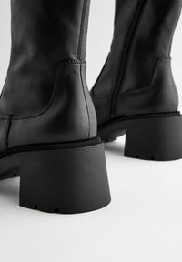 Black knee-high boots with a smooth leather finish, featuring a chunky rubber heel and side zipper detail.