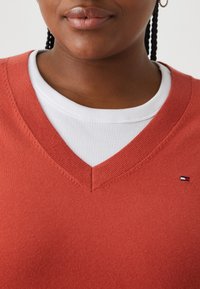 Pull en maille rouge à col en V superposé à une chemise blanche côtelée. Présente un petit logo sur le côté gauche. Texture lisse, design décontracté.