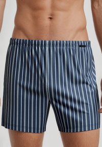 Navy blue swim shorts featuring thin white vertical stripes, an elastic waistband, and a smooth texture with a logo tag at the side.